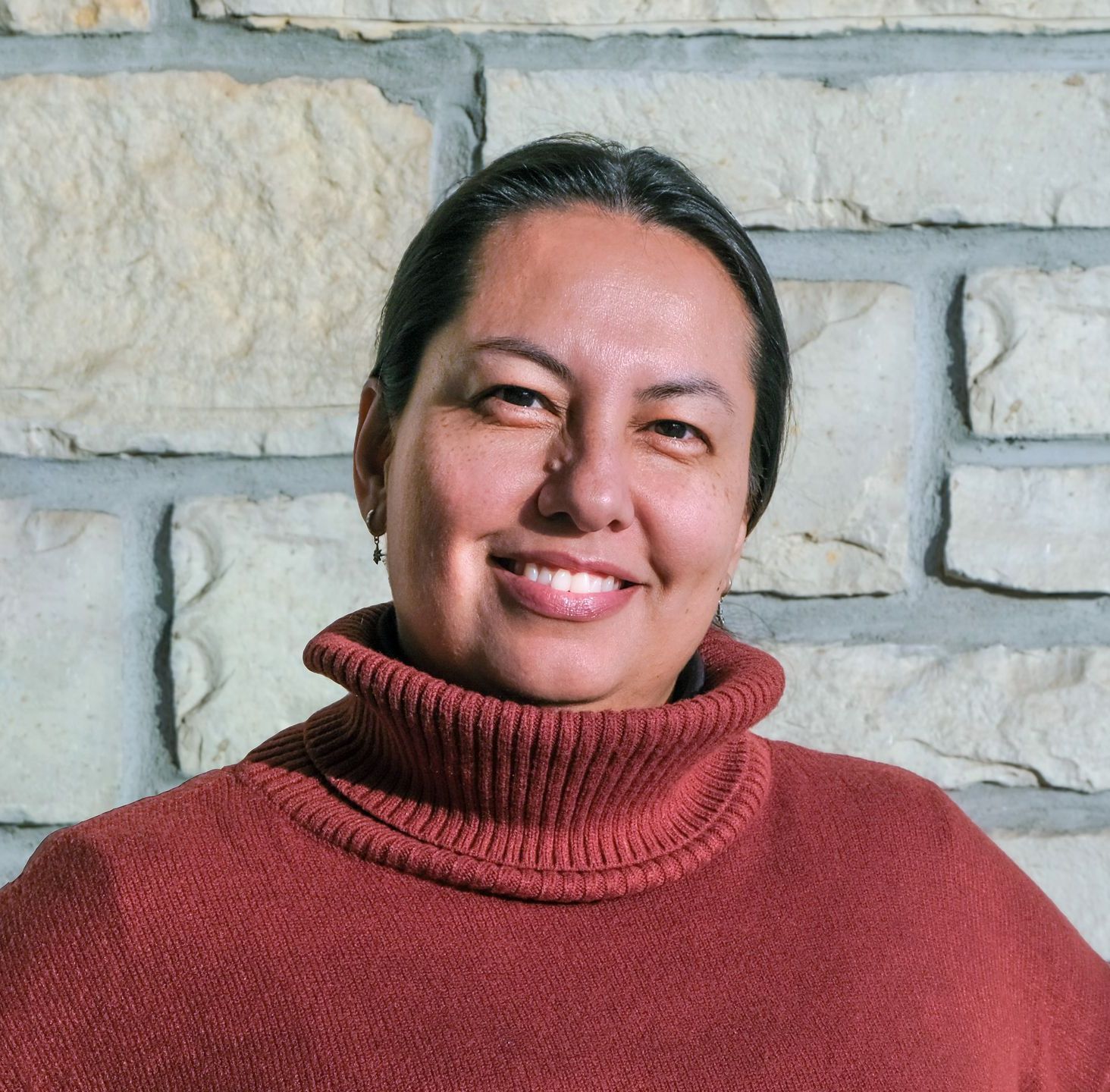A woman wearing a red sweater is smiling in front of a brick wall