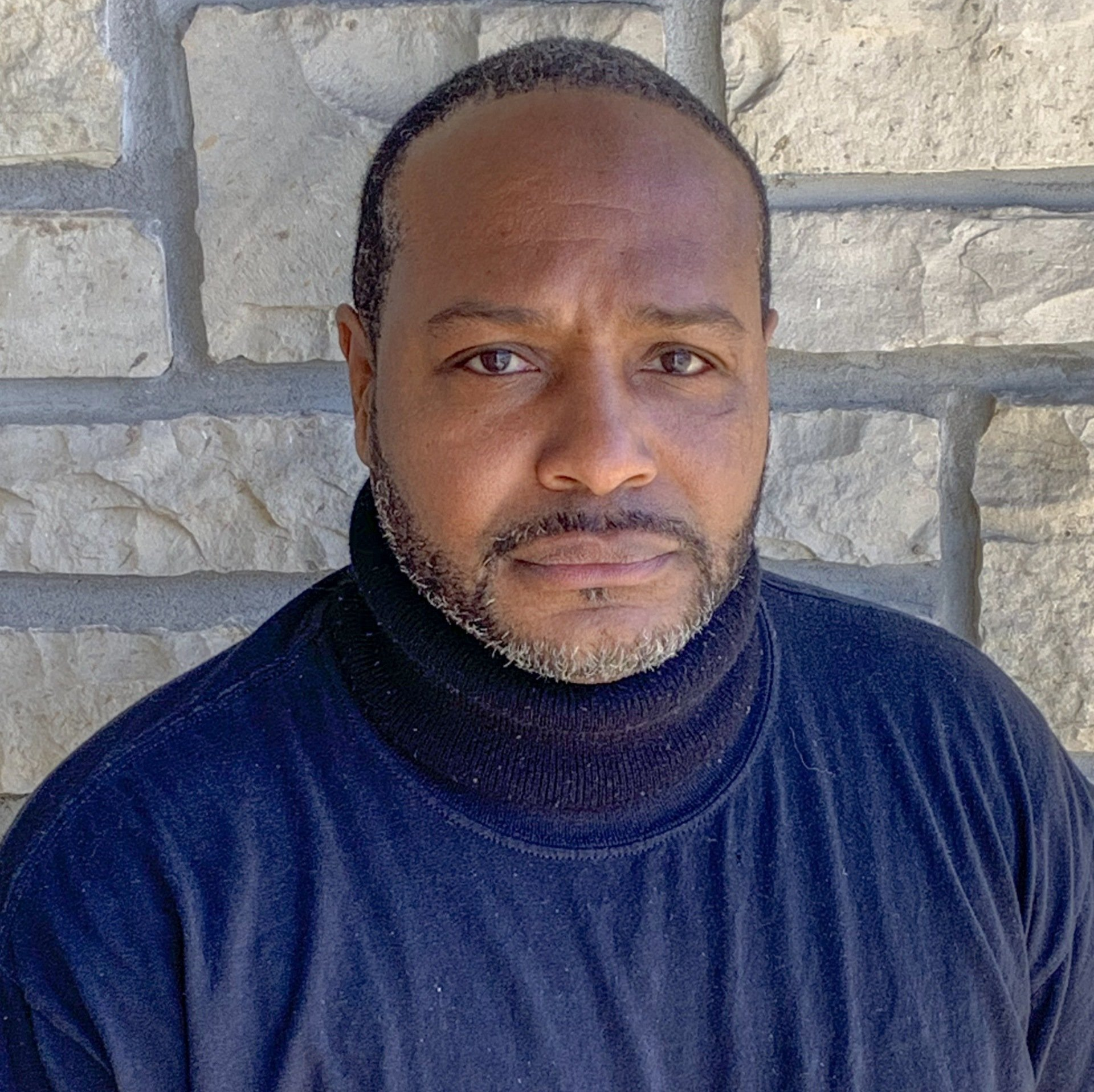 A man with a beard wearing a black turtleneck is standing in front of a brick wall.