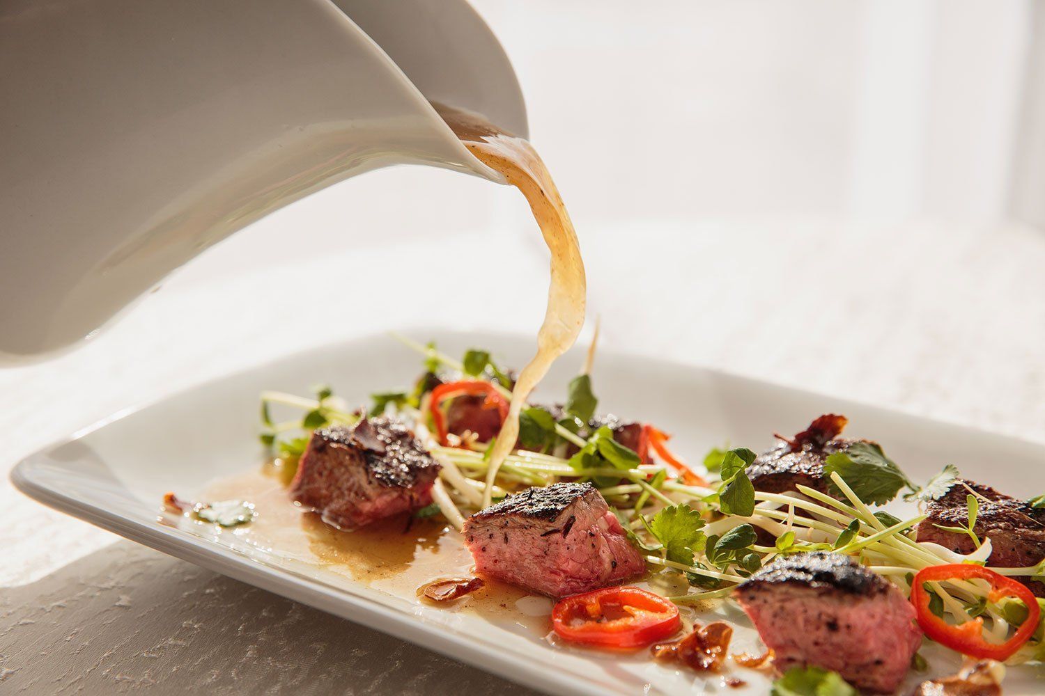 A white plate topped with meat and vegetables is being poured with sauce.