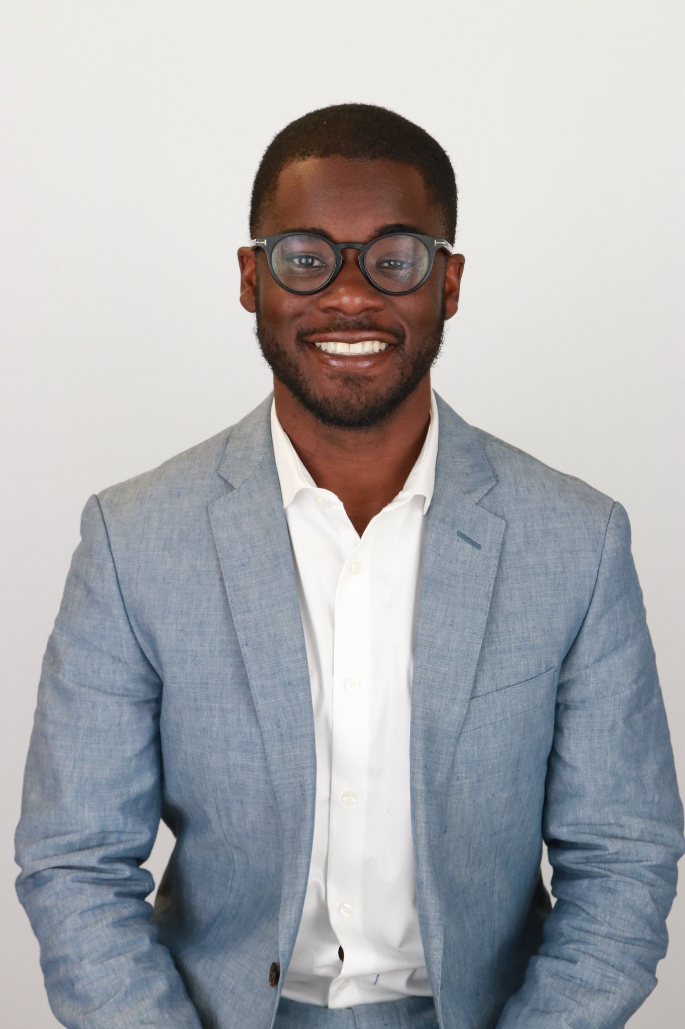 Man wearing a light blue blazer, white shirt, and glasses, smiling against a white background.
