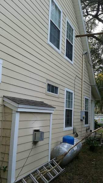 Side view of a two-story house with light yellow siding and white window frames. A small shed and propane tank sit beside the house.