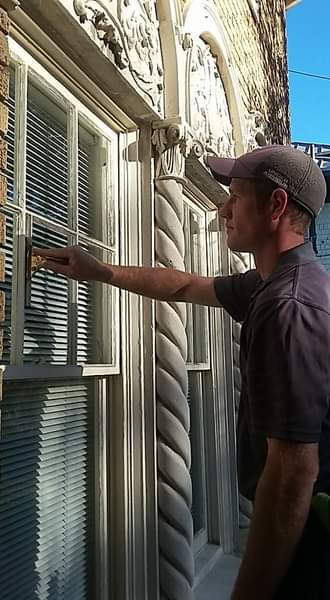 Man cleans window with a brush on a sunny day. He wears a baseball cap and stands outside a building with ornate architectural details.