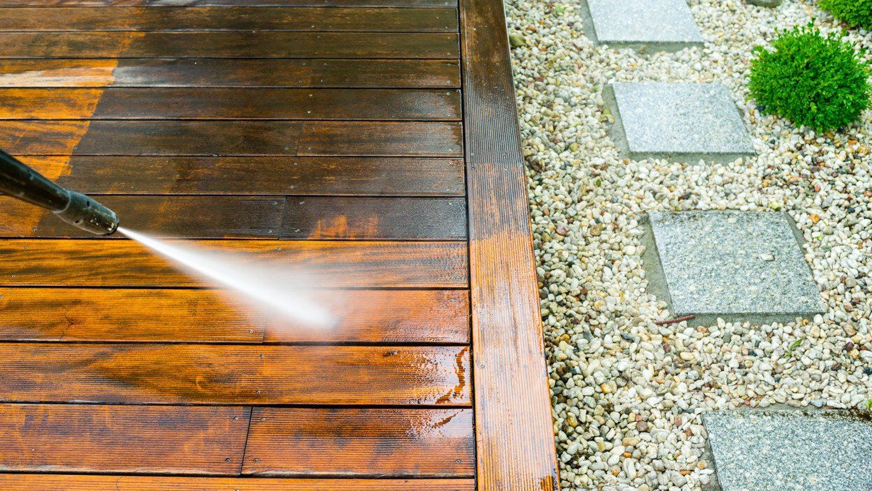 A pressure washer cleans a wooden deck, showing the contrast between cleaned and dirty sections; adjacent is a stone path.