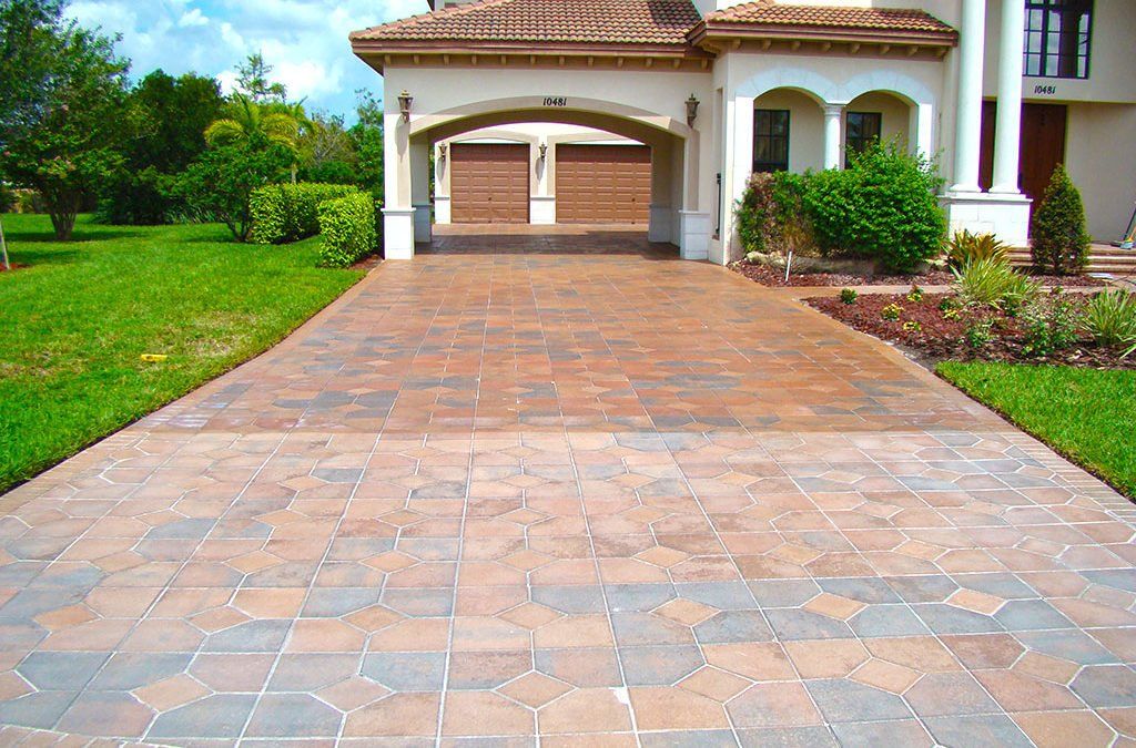 Paver driveway leading to a beige house with a two-car garage. Green lawn on either side of the driveway.