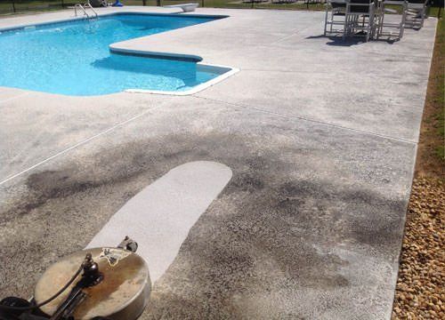A power washer cleaning a concrete pool deck, revealing a clean, light-colored path.