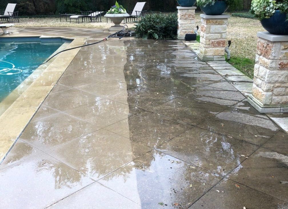 A wet stone patio, half-cleaned by a pressure washer, next to a swimming pool. The clean side is lighter and brighter.