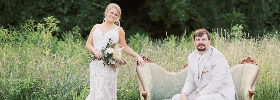 A bride and groom are posing for a picture while sitting on a couch in a field.