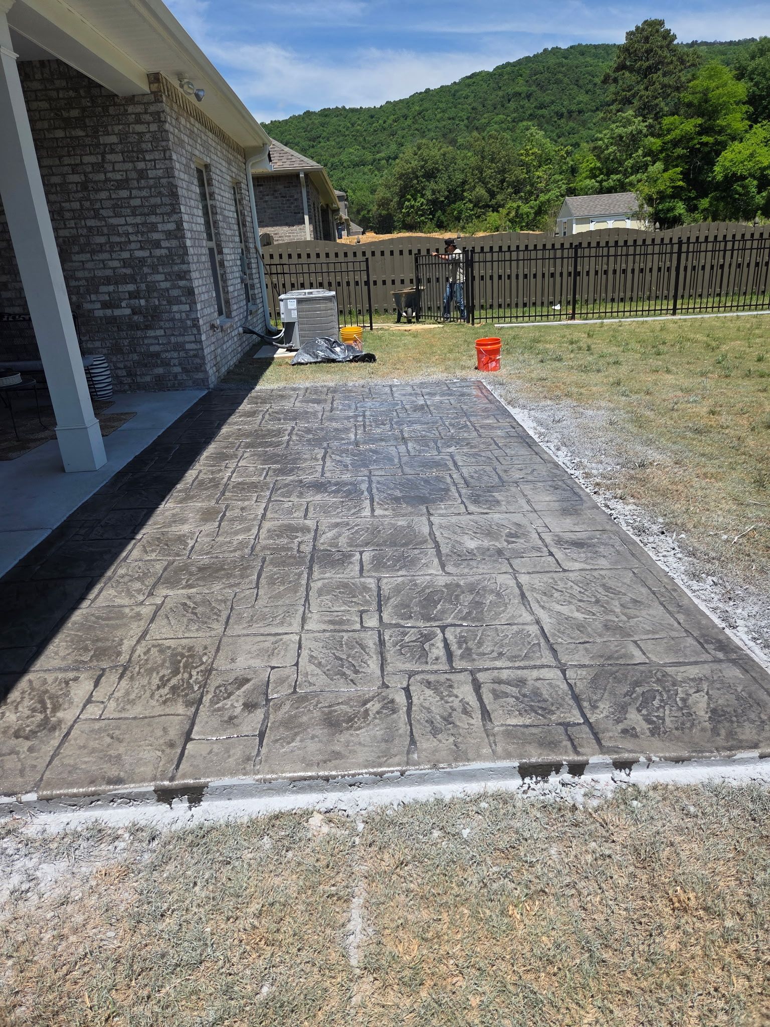 Back patio with stamped concrete, house siding, a small grill, red bucket, and fenced yard with trees beyond