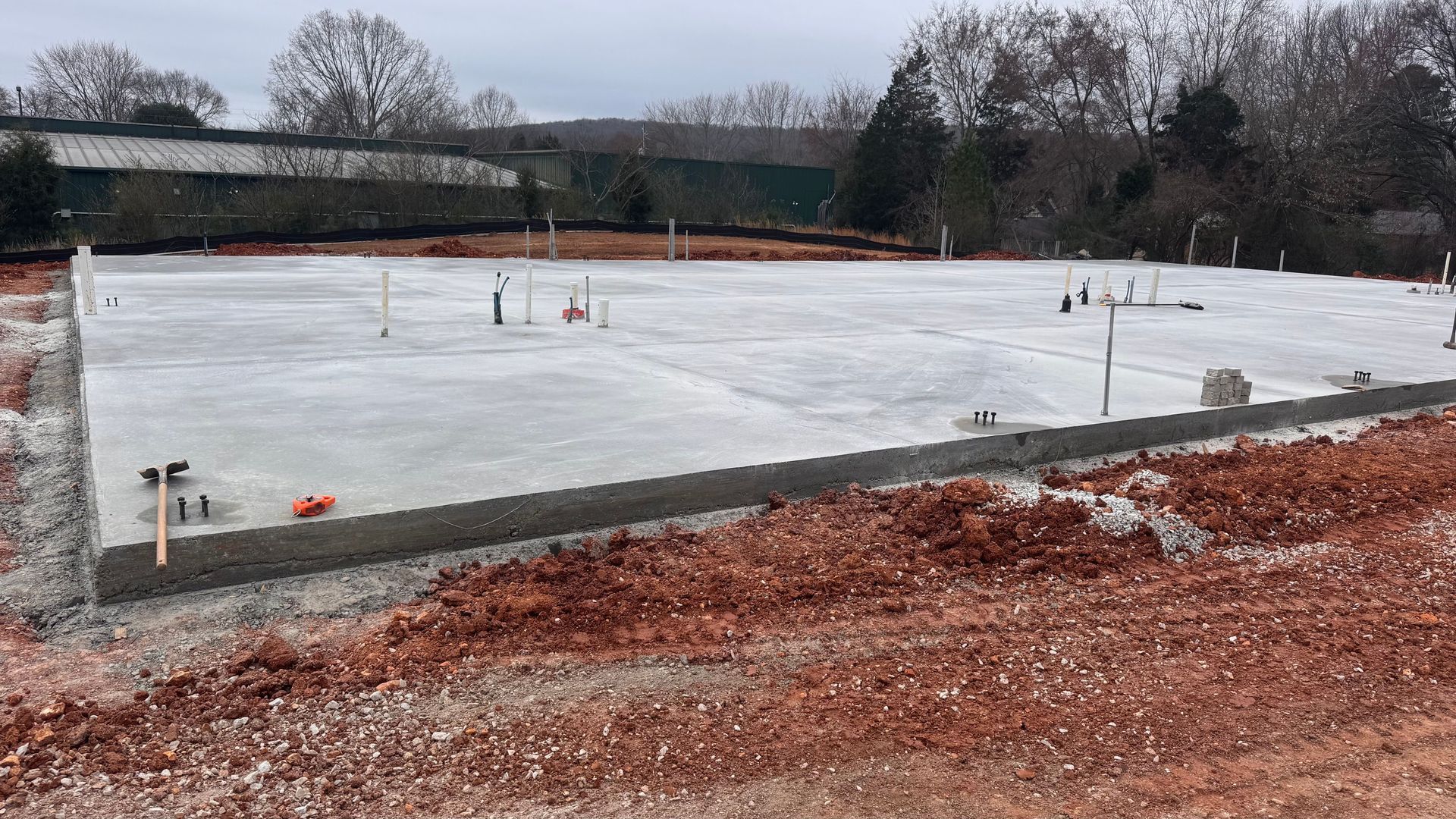 Concrete foundation slab under construction, with red dirt foreground and trees in the background