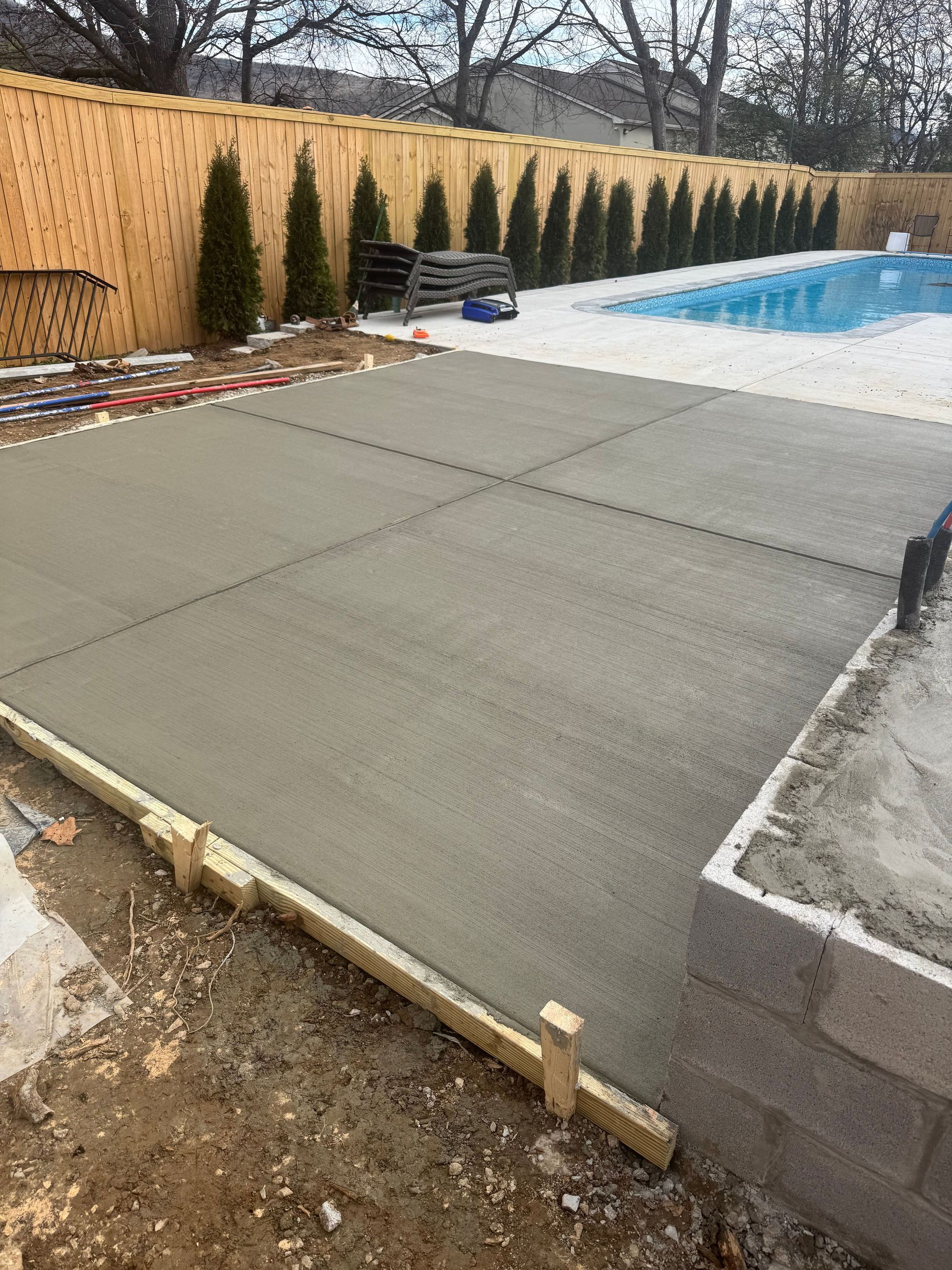 Concrete pool deck beside an in-ground pool, with a wooden fence and evergreen shrubs along the edge