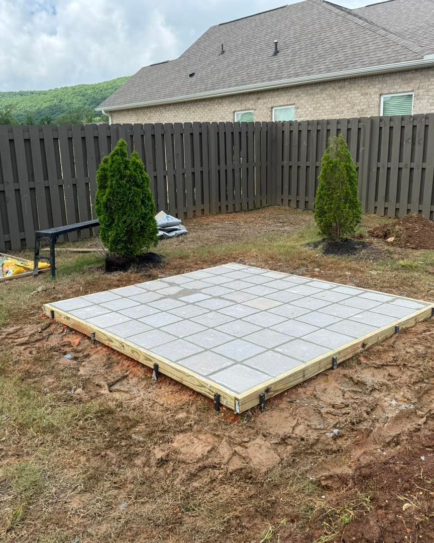 Backyard with a square tiled patio base, wooden fence, and two small evergreen shrubs.