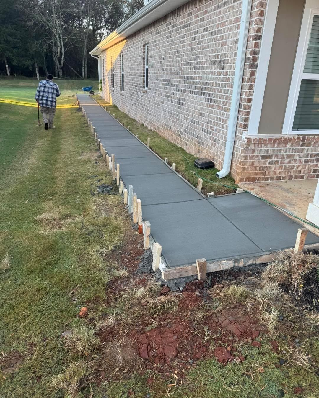 Concrete walkway under construction along a brick house, with a person walking on the grass nearby.