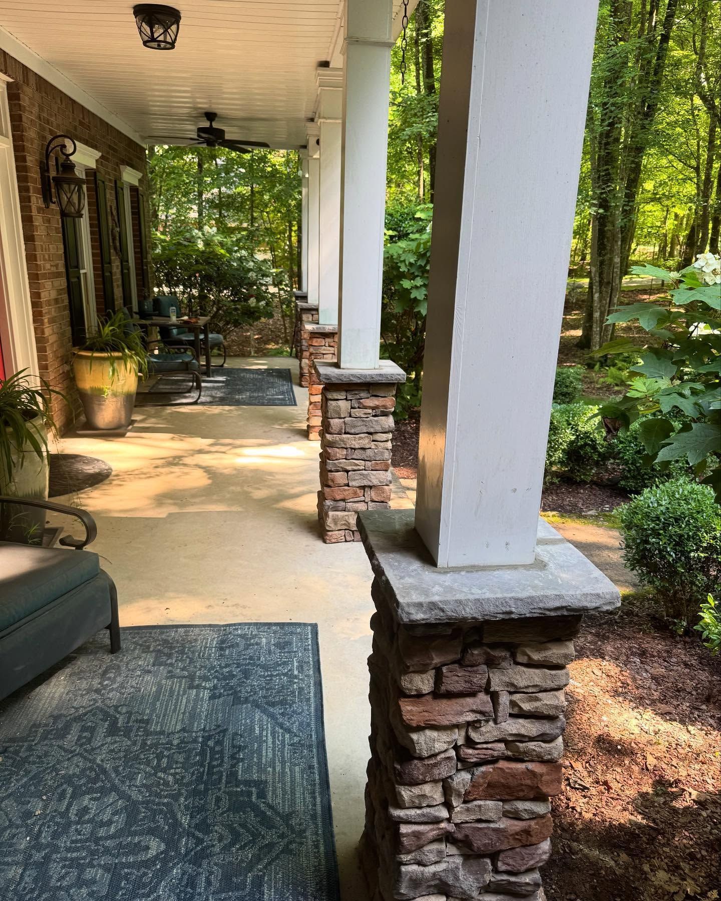 Covered front porch with stone columns, chairs, and a wooded garden view.