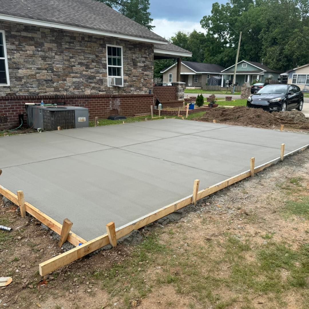Fresh concrete slab poured beside a brick house, framed by wooden forms in a backyard.