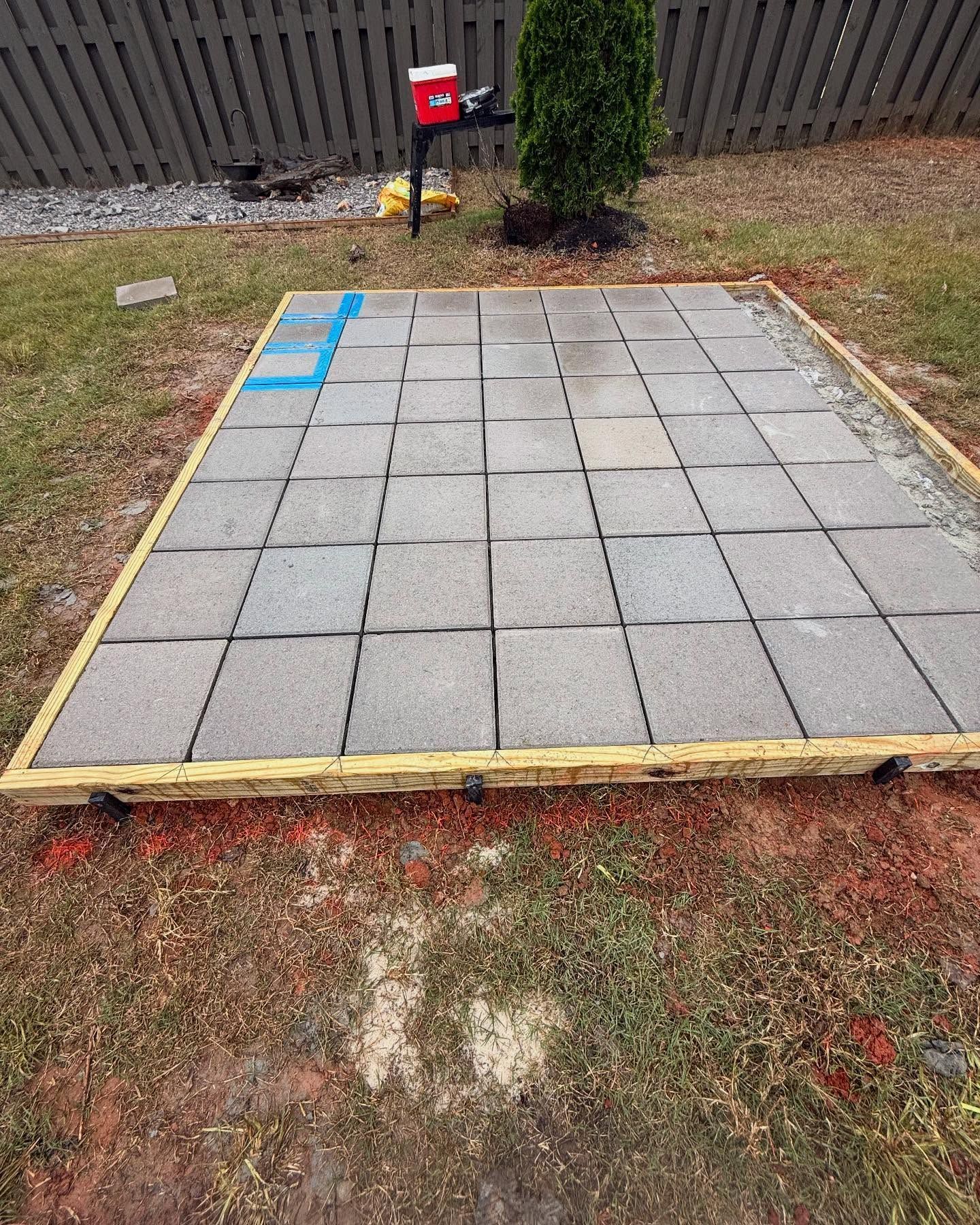Square paver patio area bordered with wood, marked with blue tape in a fenced backyard