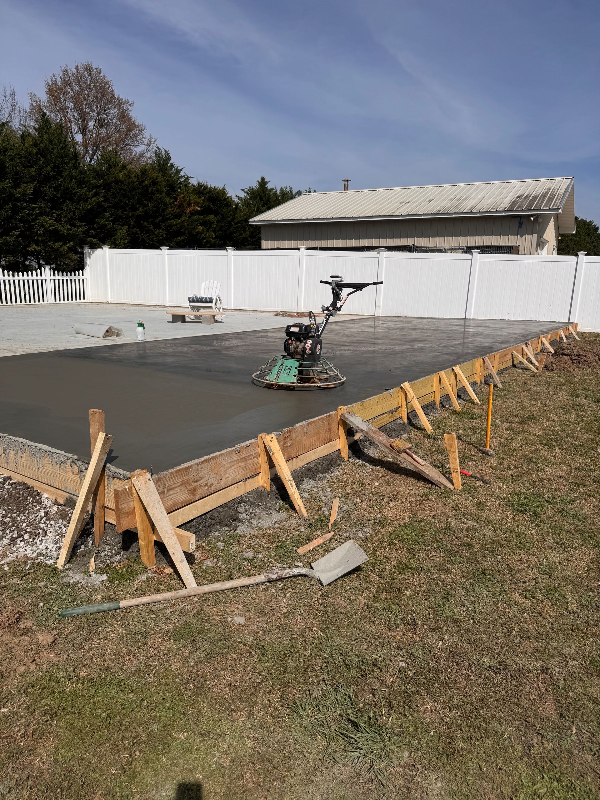 Freshly poured concrete slab with a power trowel, wooden forms, and a white fence beside a house