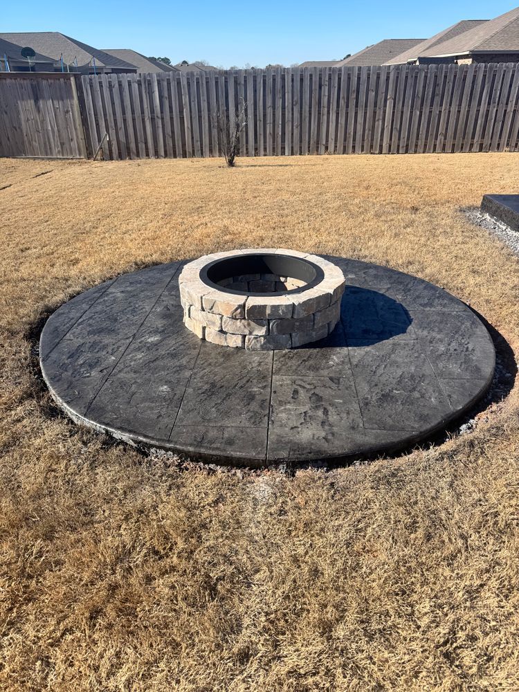 Backyard fire pit with stone ring and black circular pad on dry grass, fenced yard in background