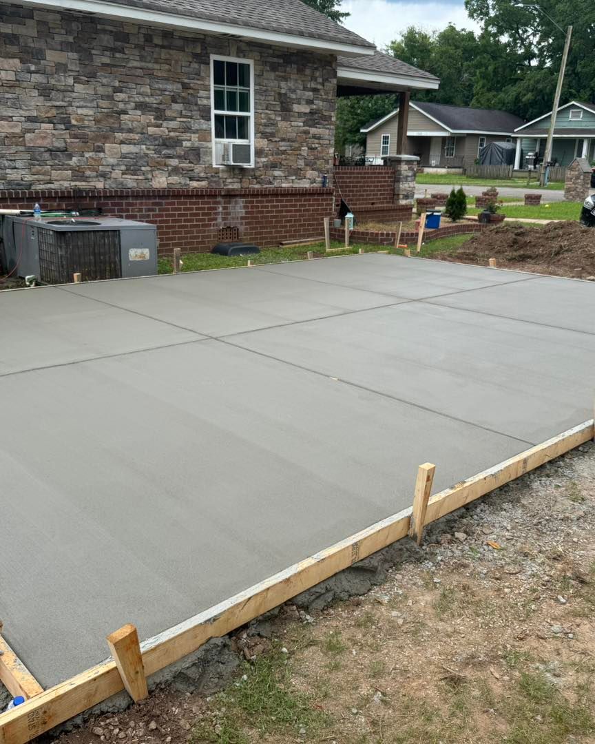 Fresh concrete patio slab beside a brick house, bordered by wooden forms and dirt yard.