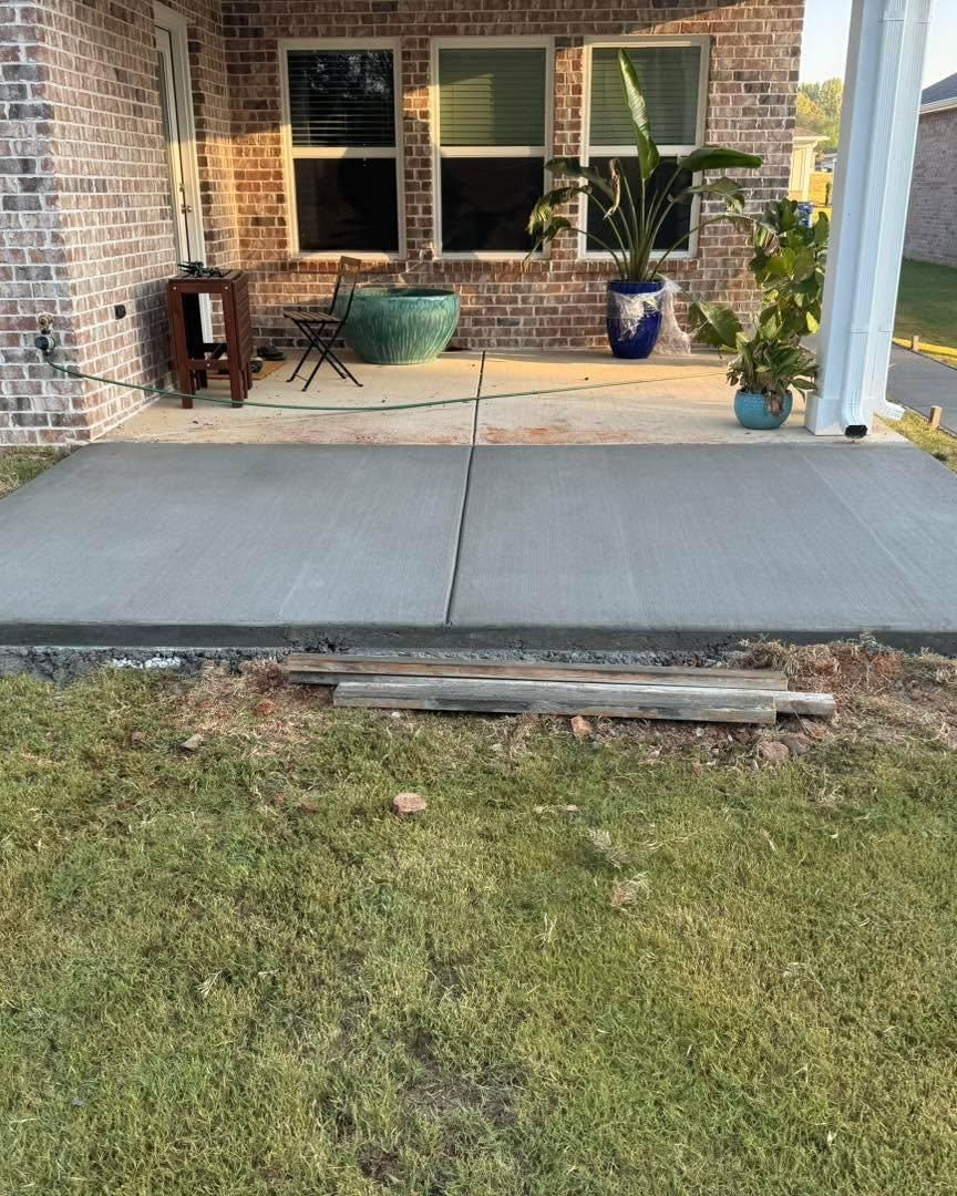 Back patio with brick wall, windows, potted plants, and a concrete slab beside a grassy yard.