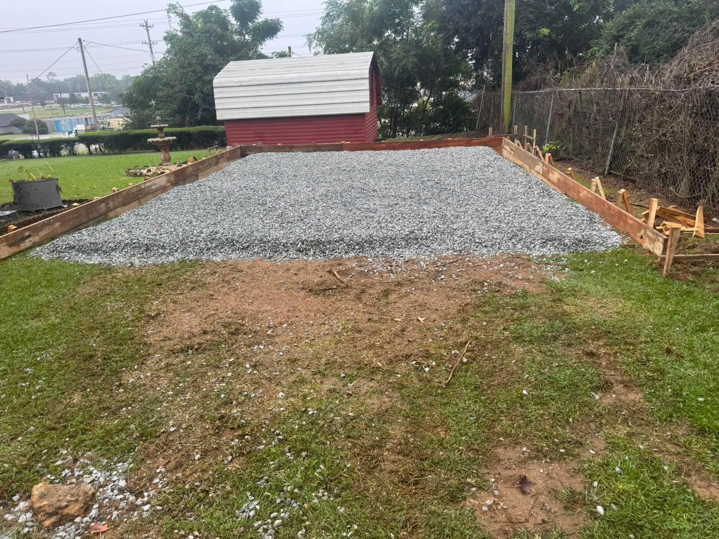 Gravel pad under construction beside a red shed, with bare dirt foreground and trees around it