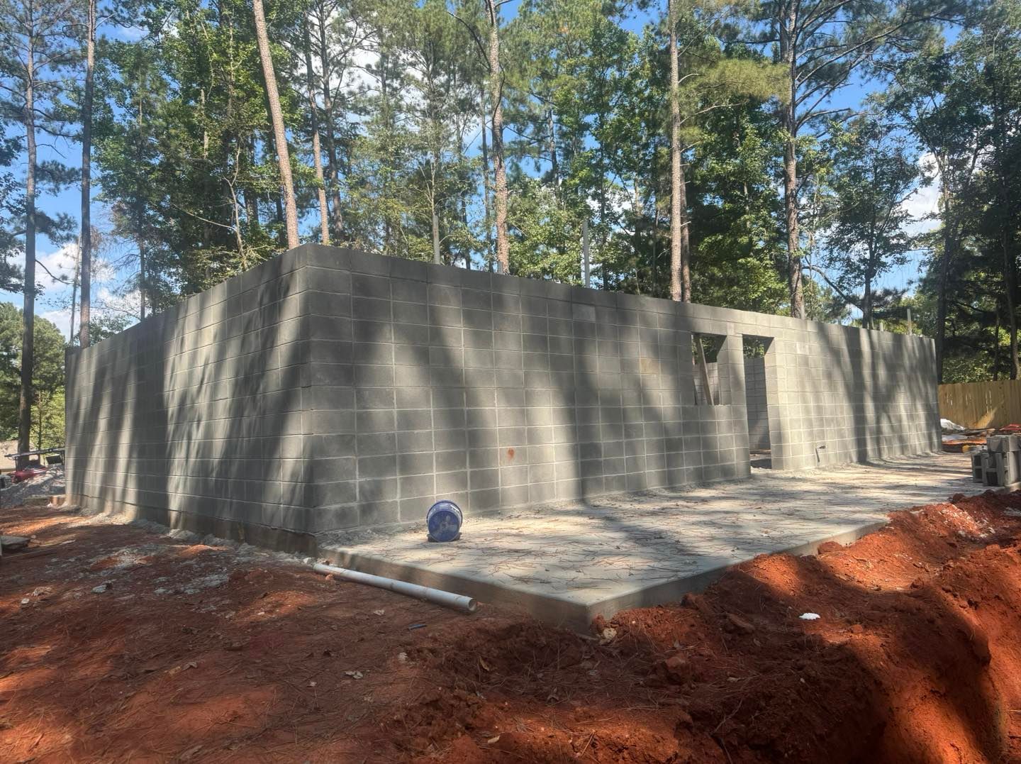Concrete foundation walls in a wooded construction site with red dirt and tall trees.