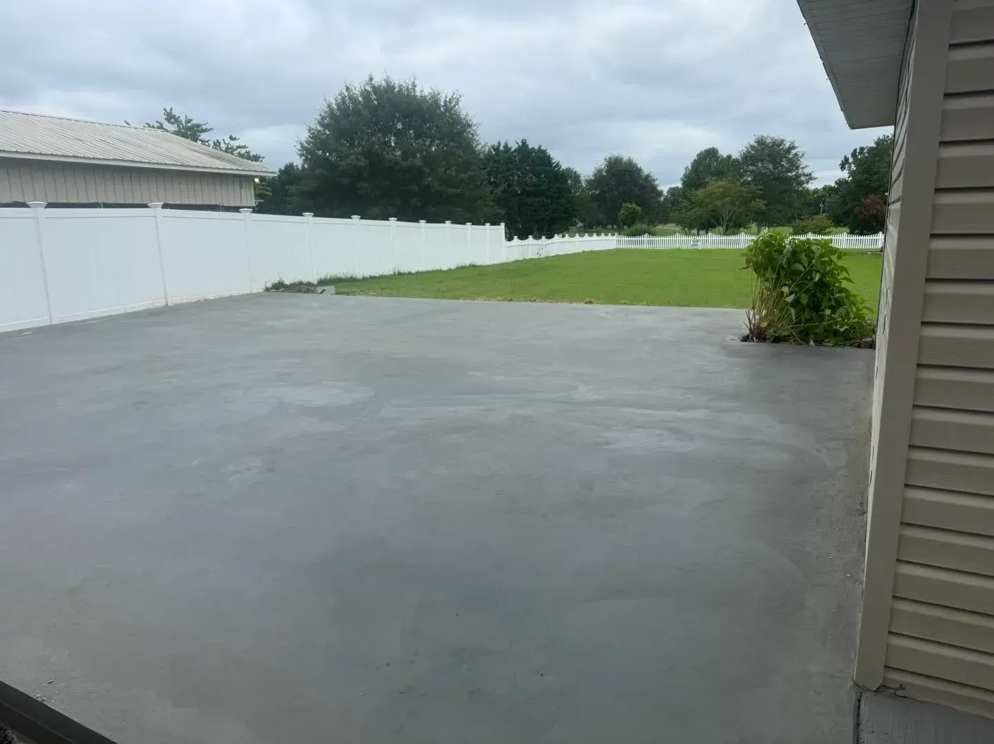 Fresh concrete patio beside a white fence, green lawn, and cloudy sky in a suburban backyard