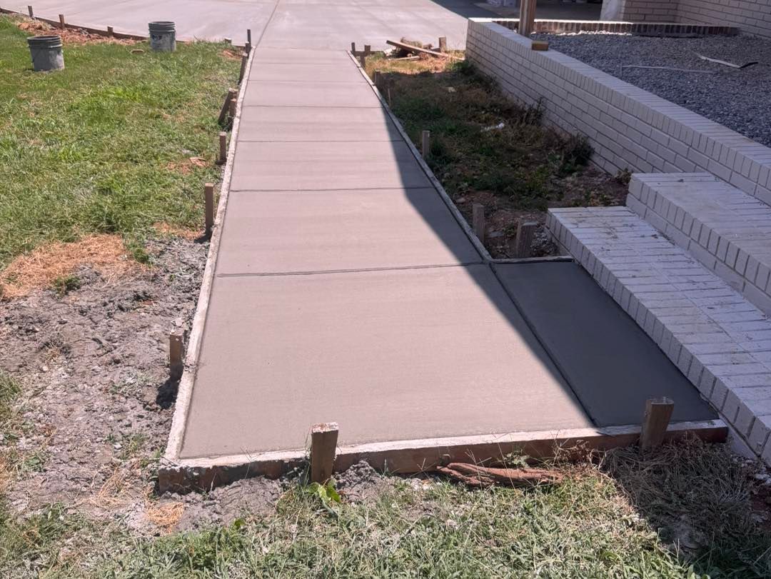Fresh concrete sidewalk beside a brick house, bordered by grass and exposed soil.