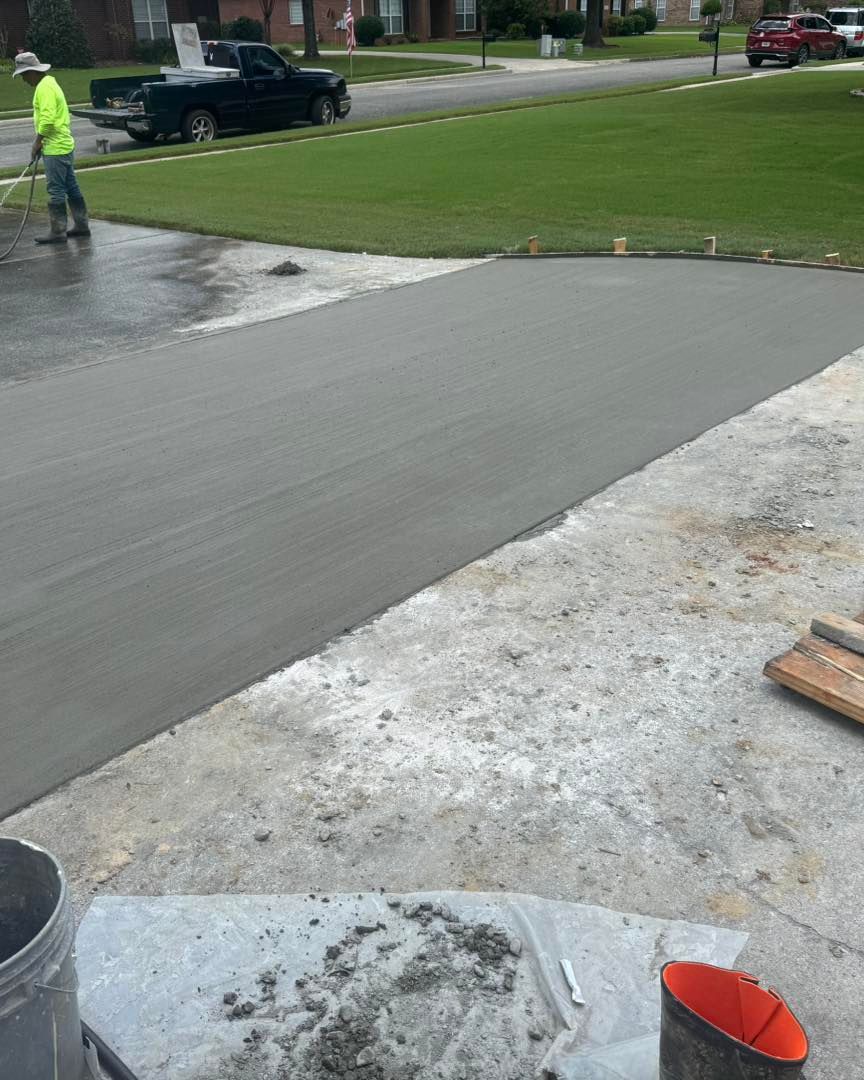 Fresh concrete driveway being smoothed, with a worker nearby on a wet residential street.
