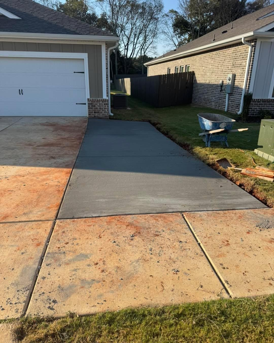 Concrete walkway beside a garage, with a fresh dark-gray section leading to a fenced backyard.
