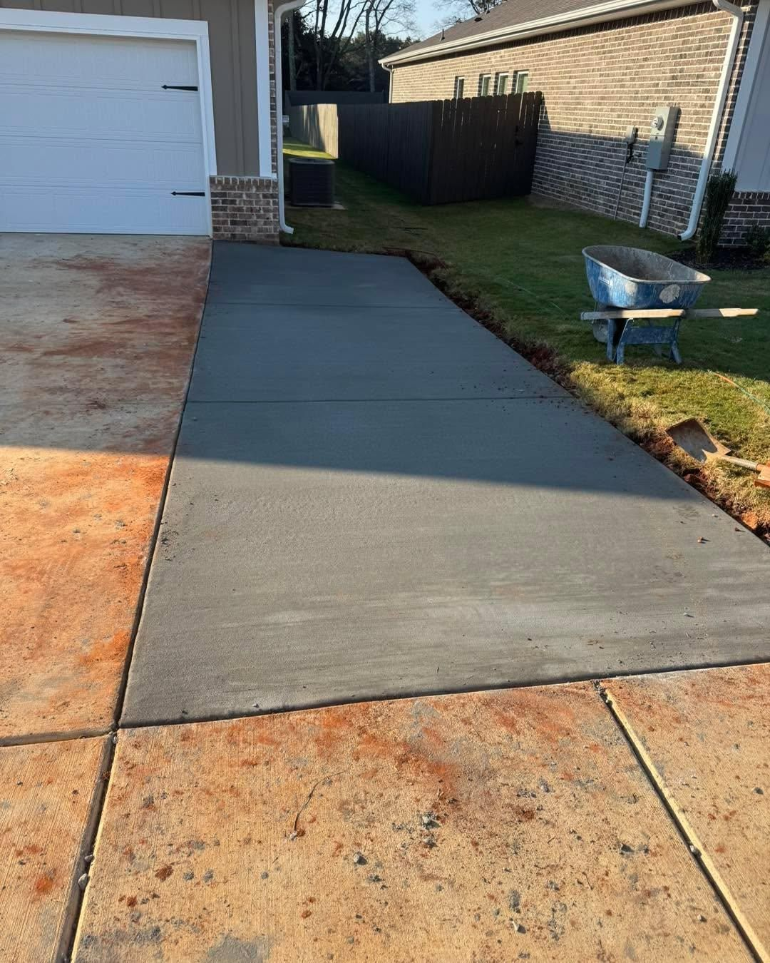 Freshly poured dark gray concrete driveway beside a garage and lawn, with a wheelbarrow on the grass.