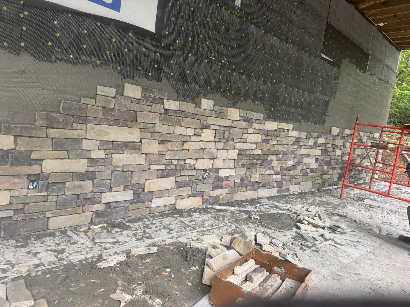 Snowy construction site with a stone wall, orange safety barrier, and a broken cardboard box on the ground