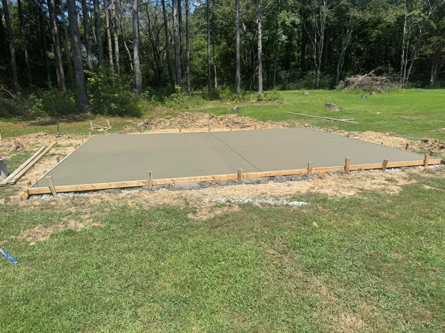 Freshly poured concrete slab on a wooded construction site, bordered by grass and gravel.