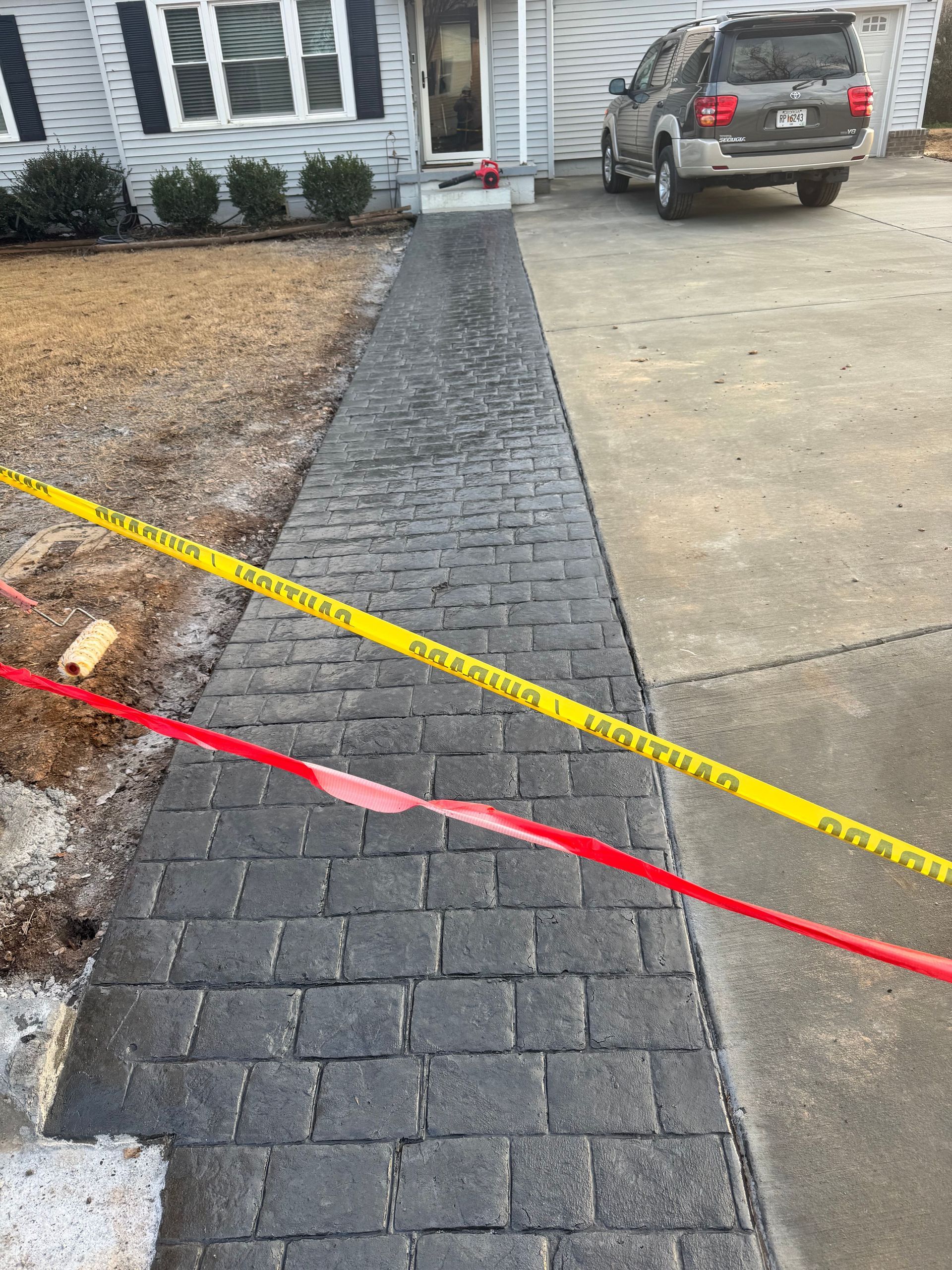 Driveway with a newly paved dark brick walkway and yellow caution tape leading to a house and parked SUV.