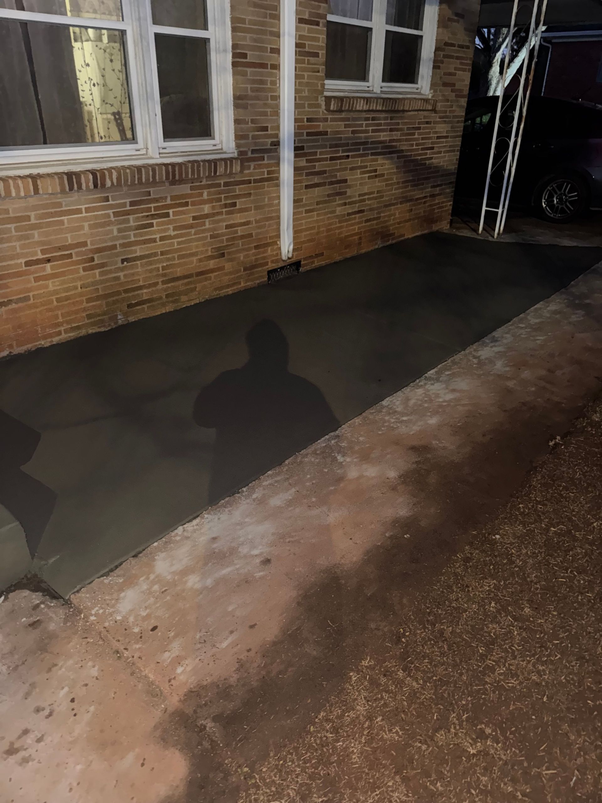Wet concrete walkway beside a brick house at night, with a shadow on the pavement.