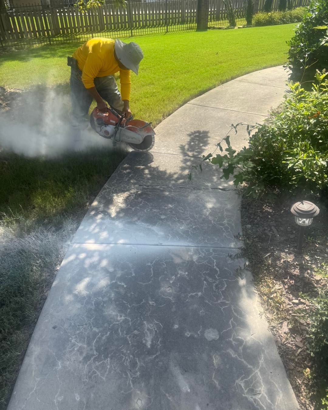 Worker cutting concrete sidewalk with a saw, creating dust in a sunny yard