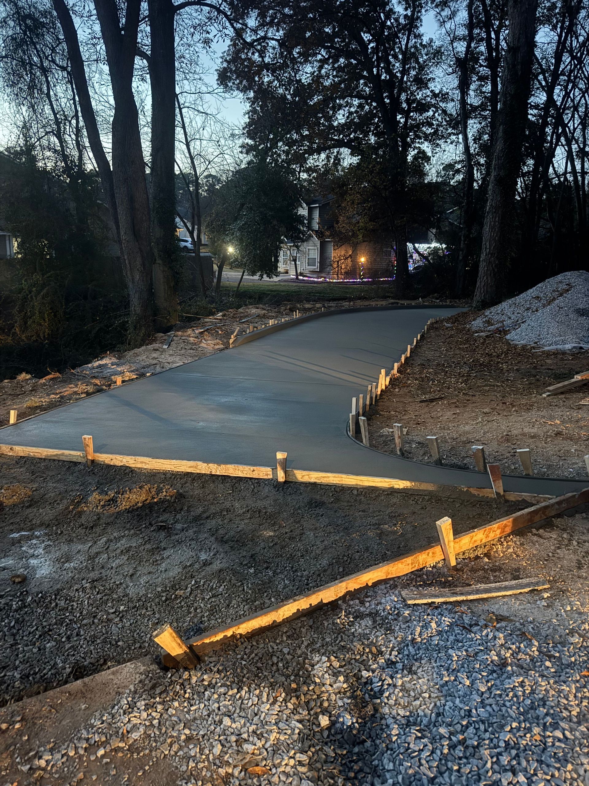 Curved concrete path under construction at dusk, with wooden stakes and dirt borders in a wooded area
