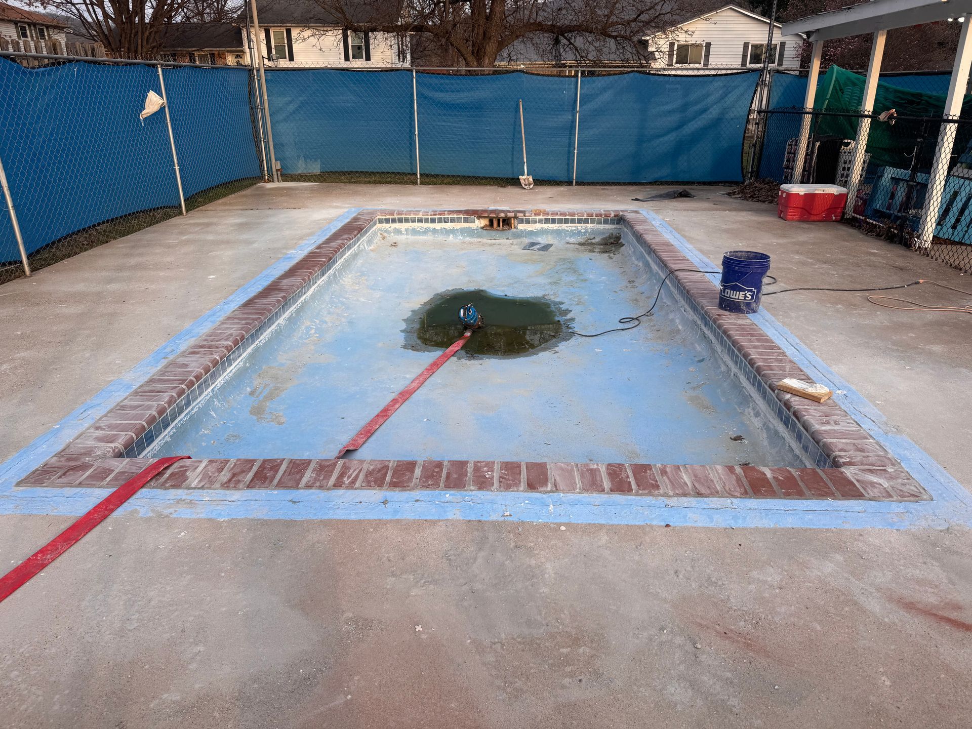 Shallow empty pool with cracked concrete, blue fencing, and a blue bucket nearby