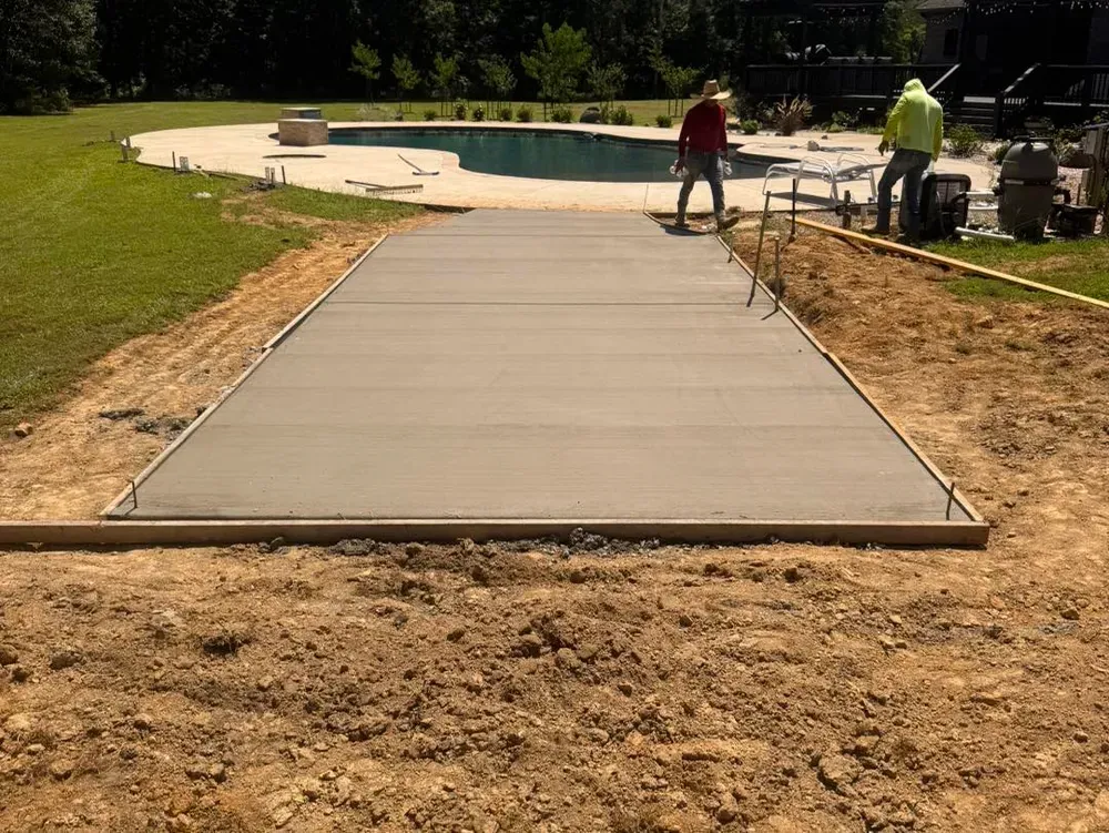 Workers install a concrete pool deck beside an outdoor swimming pool and grassy lawn.