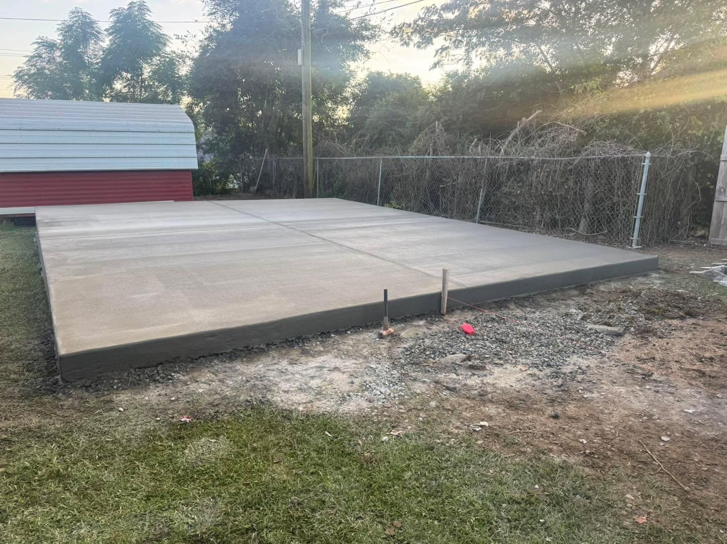 Concrete slab foundation in a backyard beside a shed, with trees and fencing in the background.