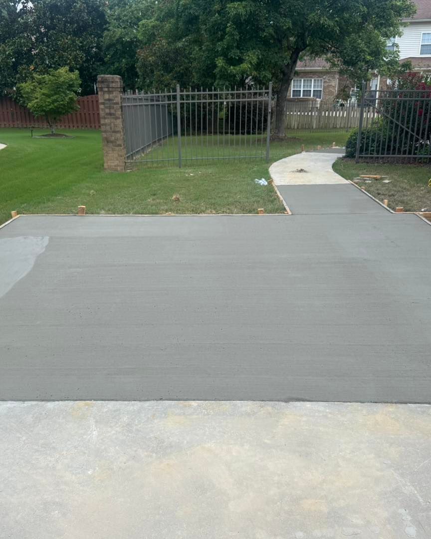 Concrete driveway leading to a sidewalk, with grass, trees, and black metal fencing in a residential area