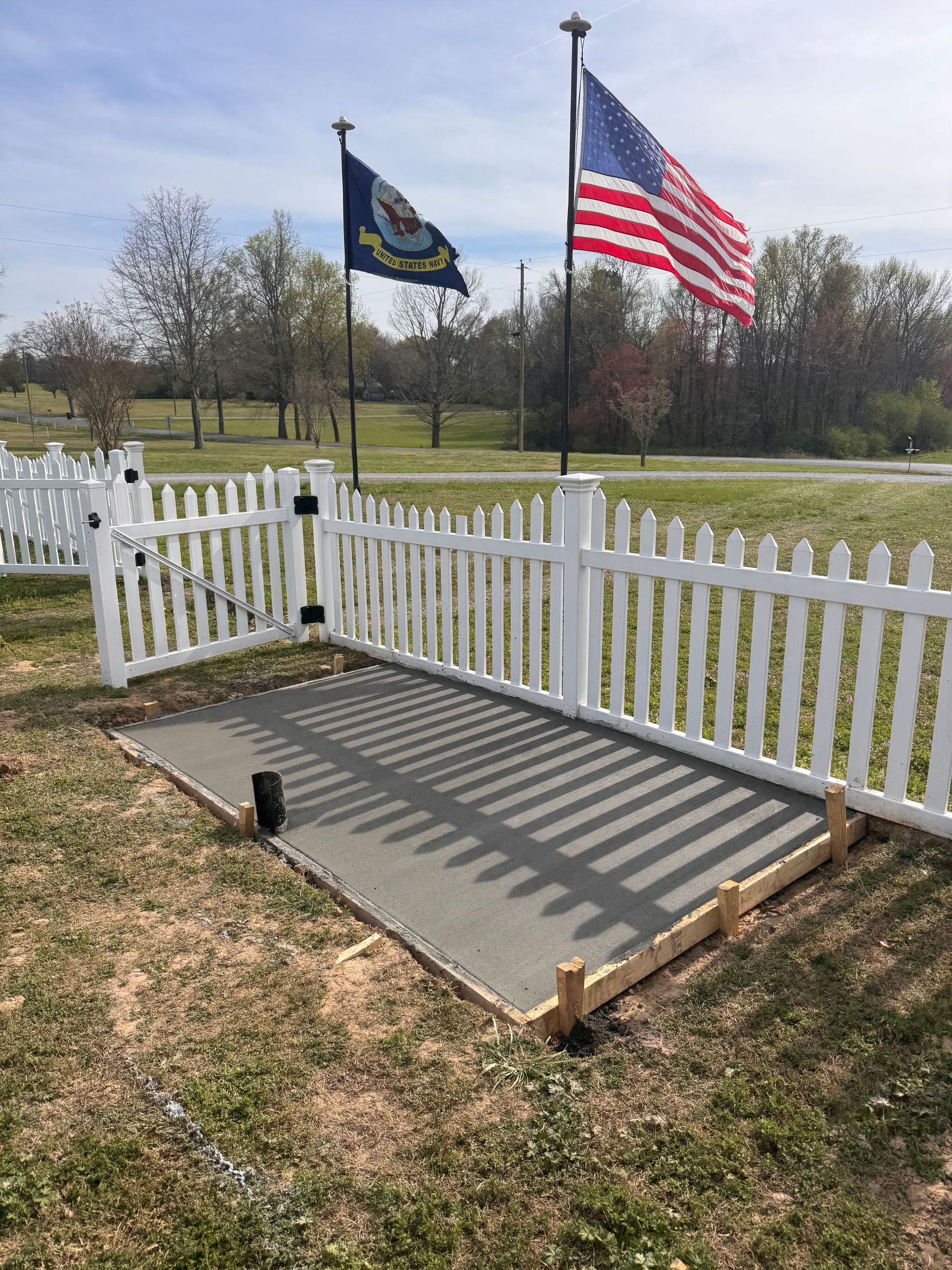 White fence and gate around a small ramp with U.S. and state flags in a grassy yard.