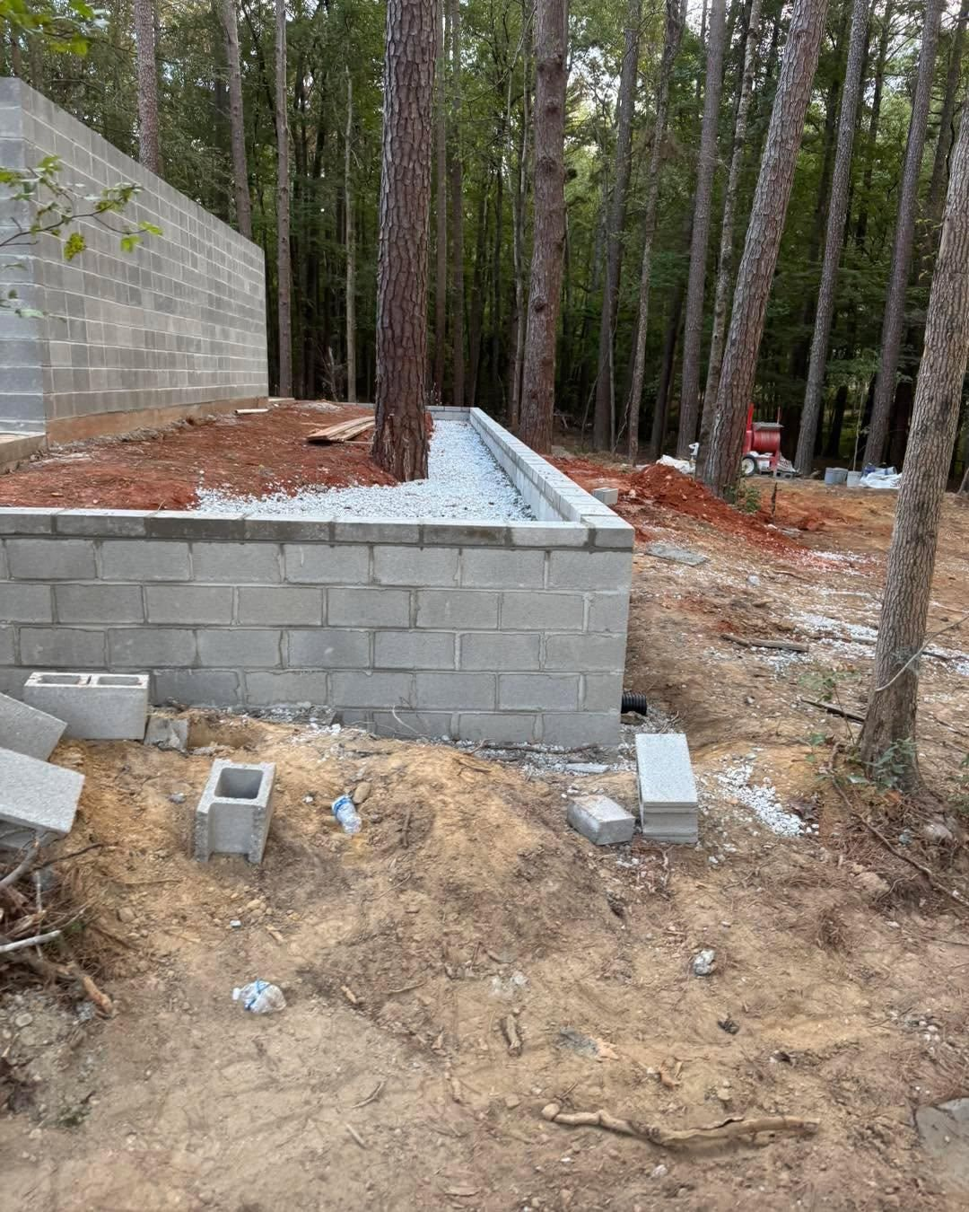 Unfinished cinder block foundation wall in a wooded construction site with dirt and gravel.