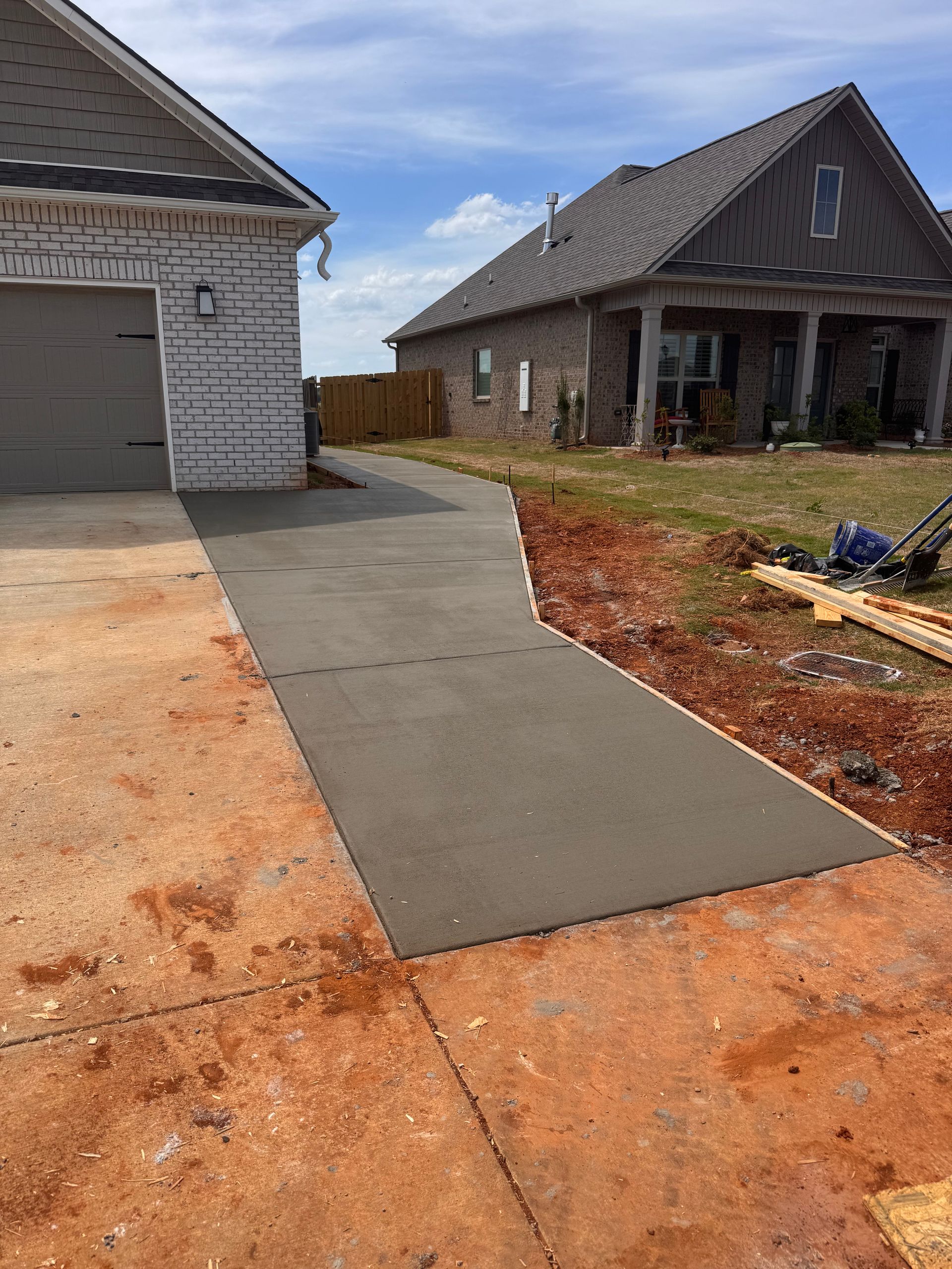 Concrete driveway under construction beside a house, with unfinished dirt edges and a second home in the background.