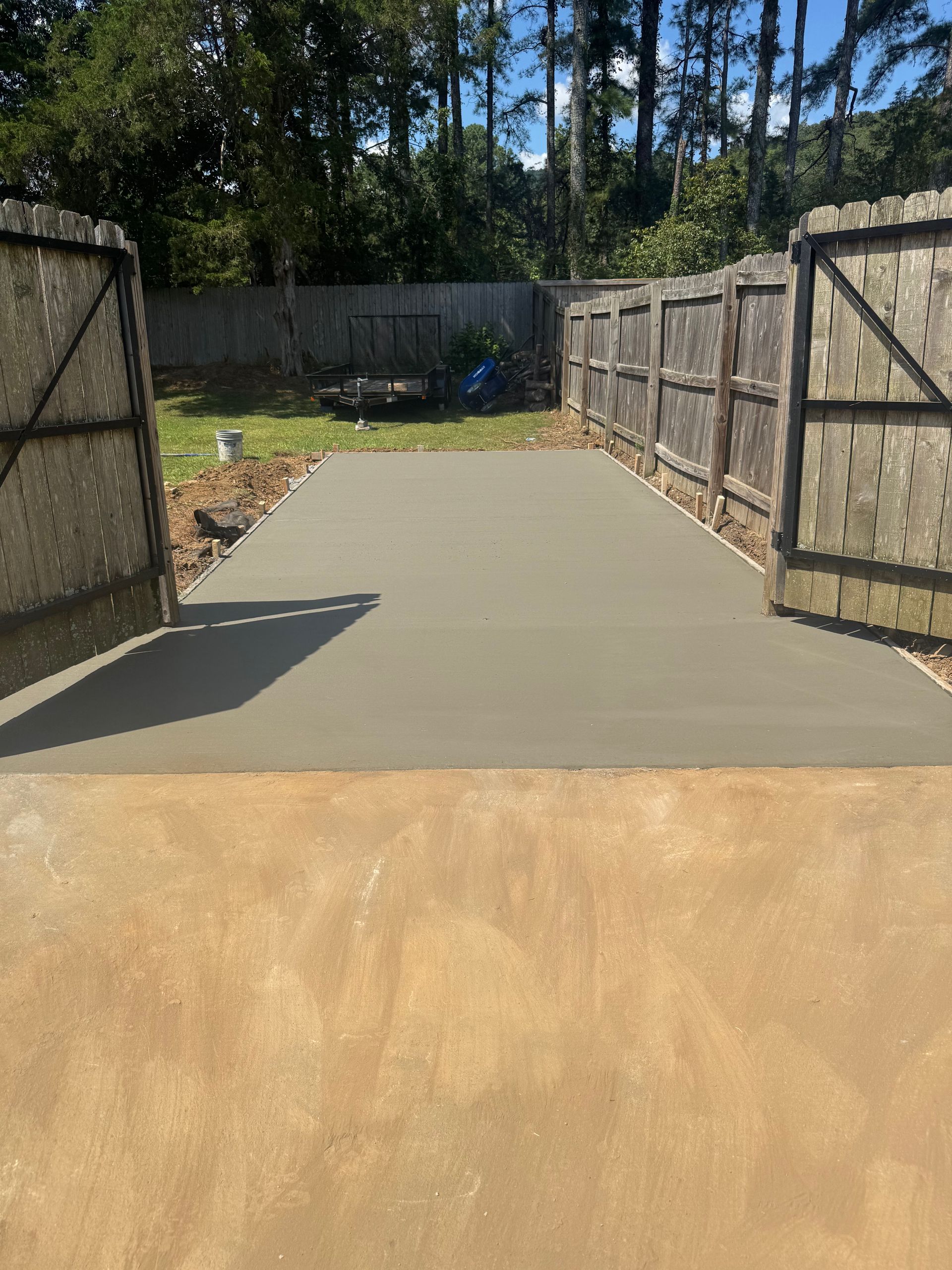 Fresh concrete slab between open wooden fence gates in a backyard under bright sunlight