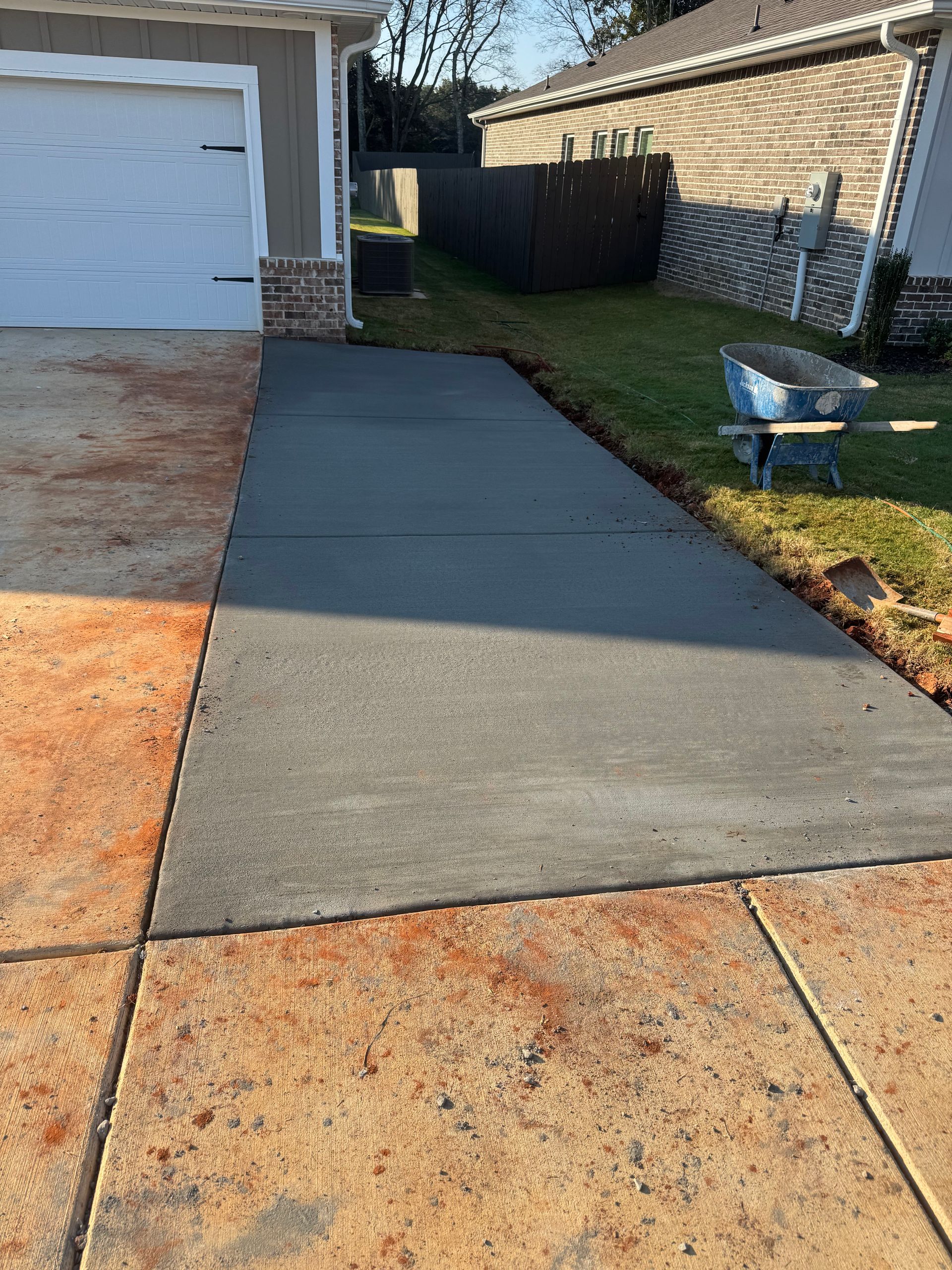 Freshly poured gray concrete driveway apron beside a garage and brick house, bordered by grass and a wheelbarrow