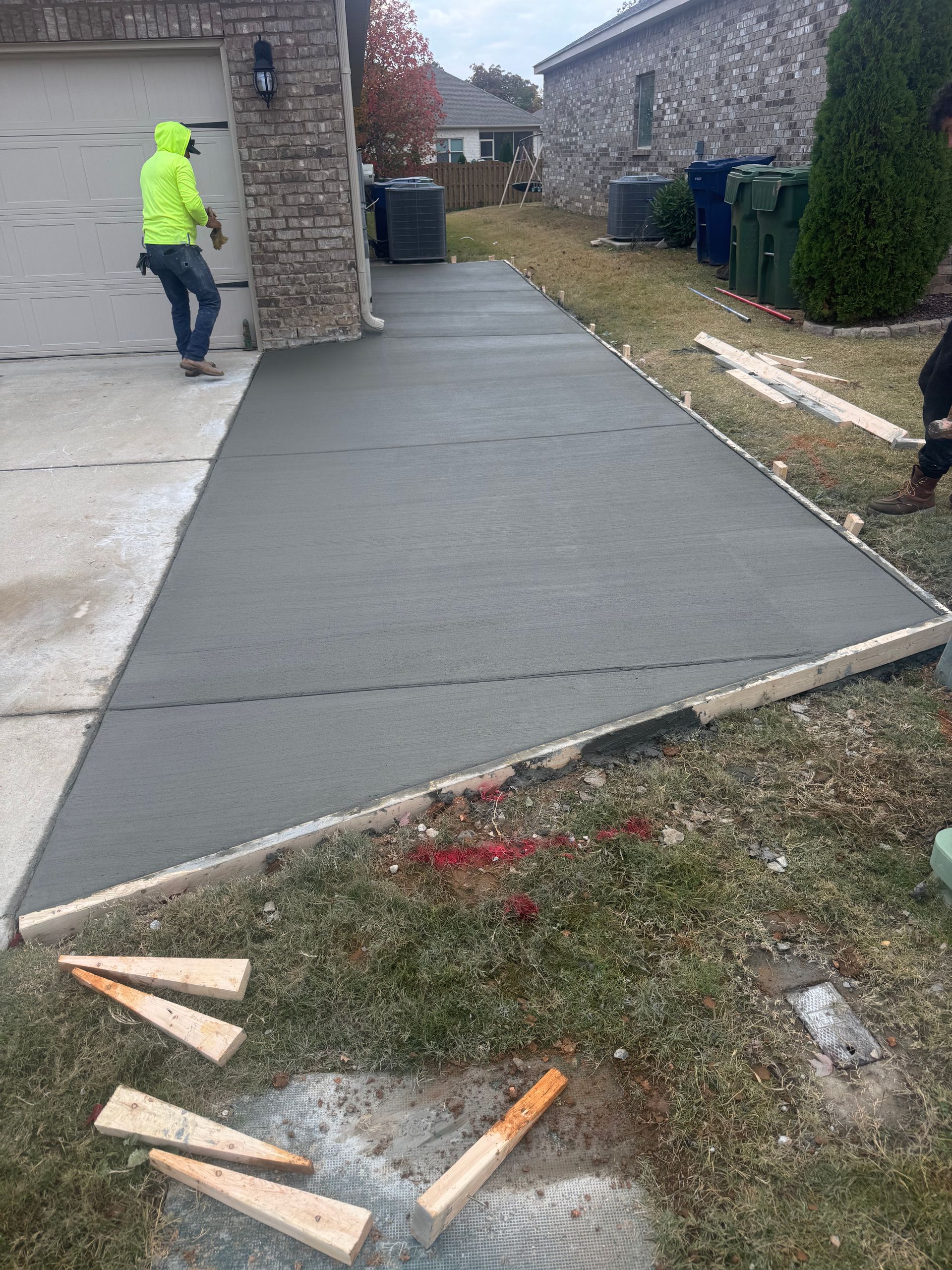 Freshly poured concrete walkway beside a house, with a worker in a neon jacket nearby.