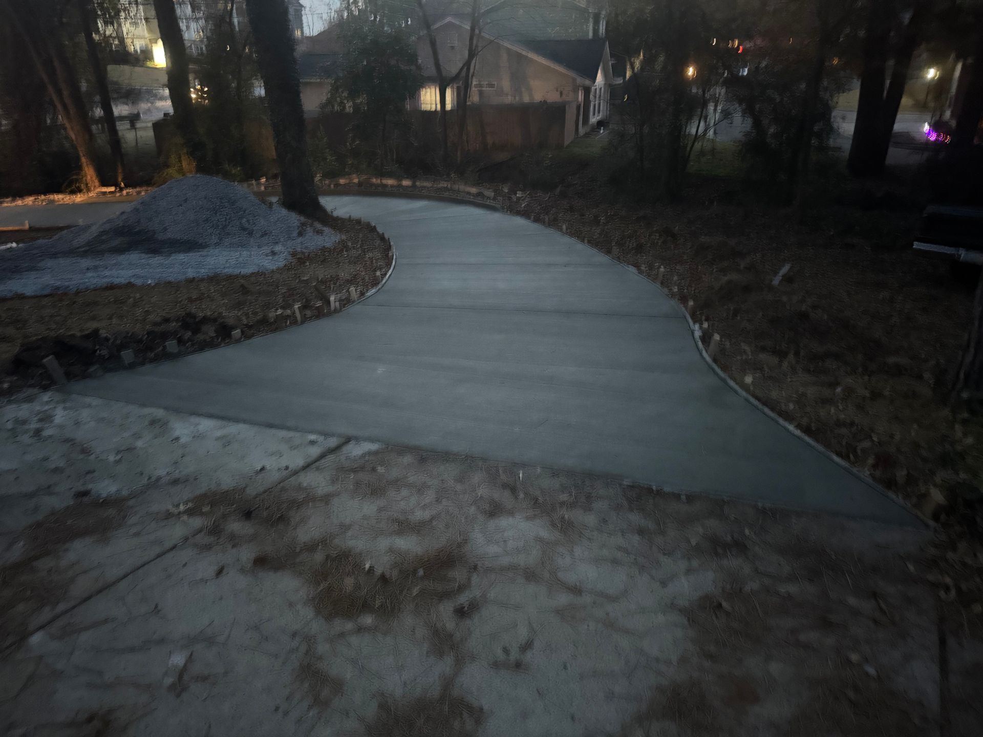 Curved concrete sidewalk at dusk beside bare trees and a small house, with patches of snow nearby