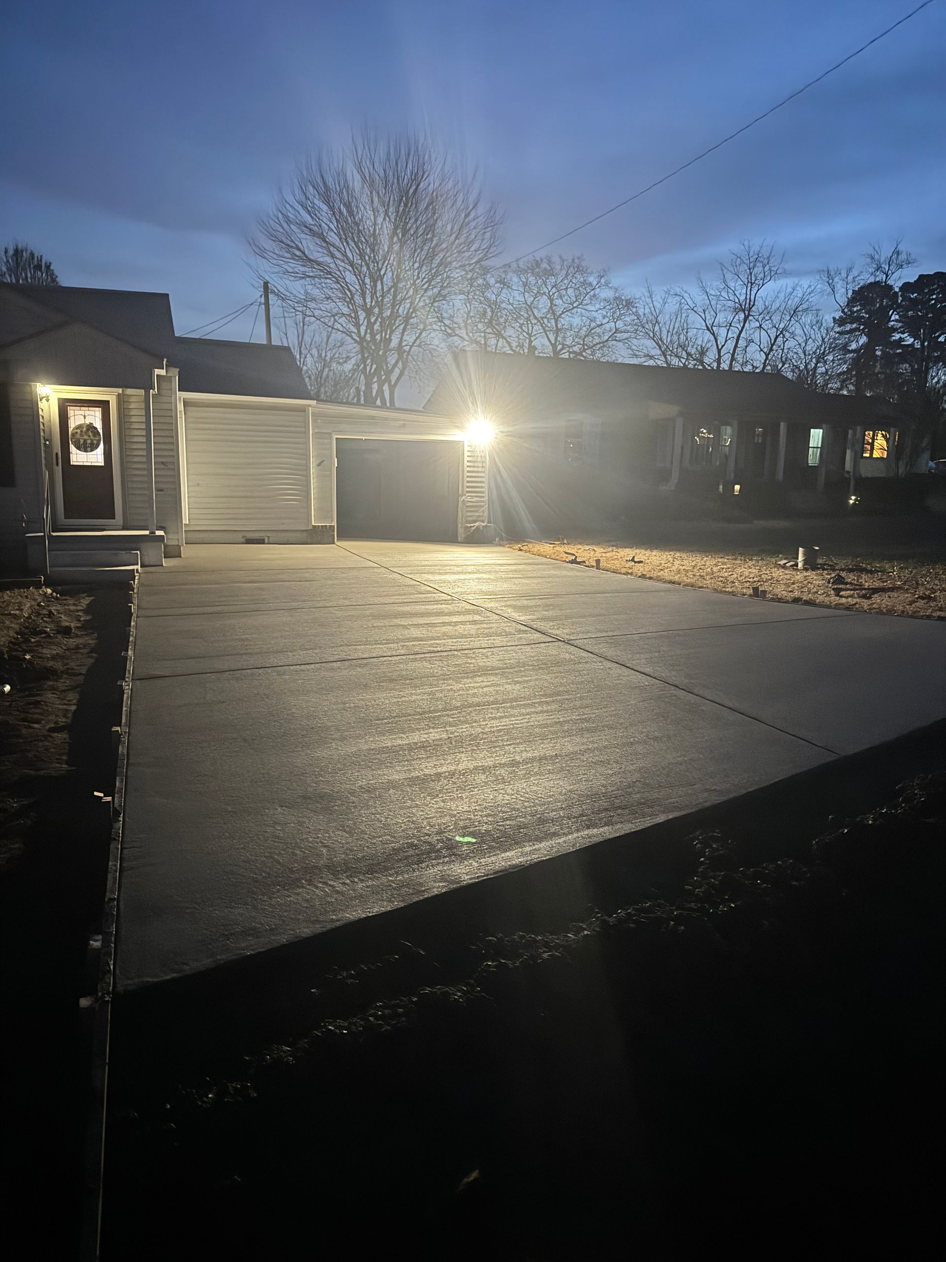 Dim residential driveway at dusk with a lit garage and bare trees silhouetted against a blue sky