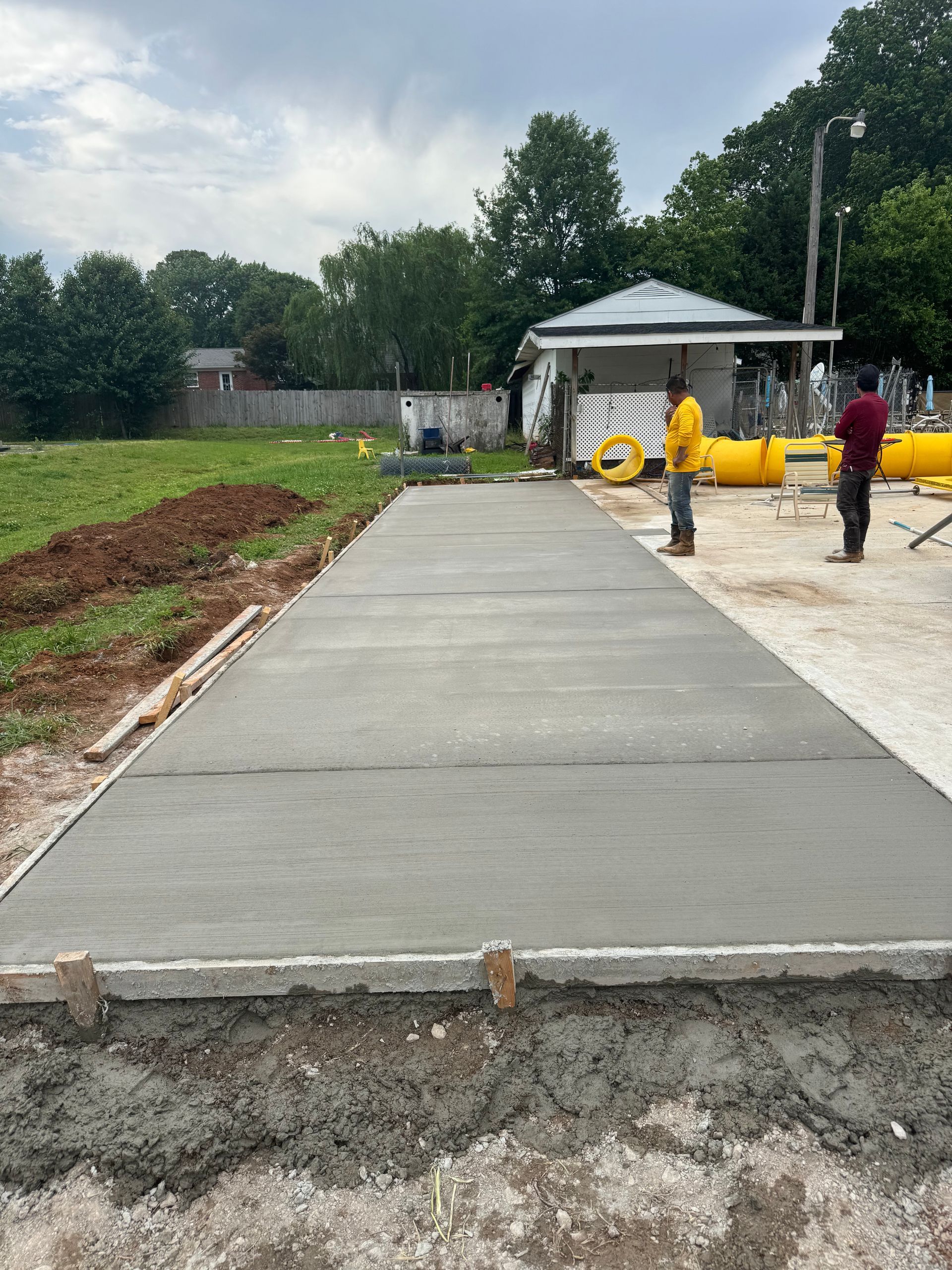 Fresh concrete driveway under construction with workers, gravel, and a small shelter in the background