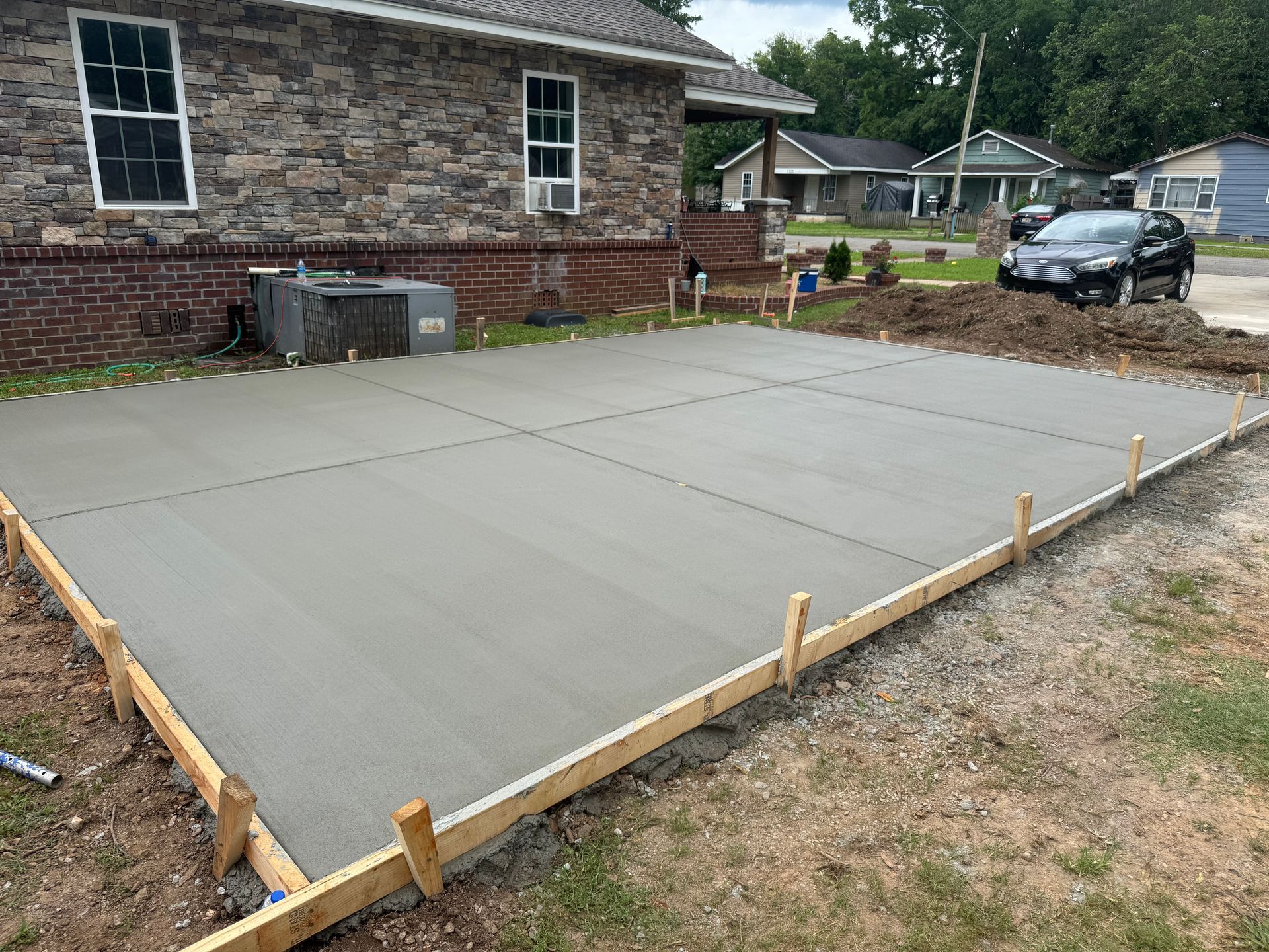 Freshly poured concrete driveway foundation beside a brick house, with wooden forms and a parked car nearby